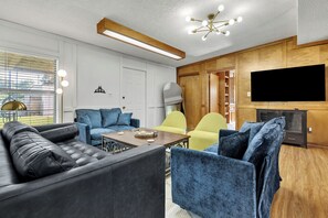 Ein modernes Wohnzimmer mit einem schwarzen Ledersofa, blauen und gelben Sesseln, einem Holztisch und einem an der Wand befestigten Flachbildfernseher.