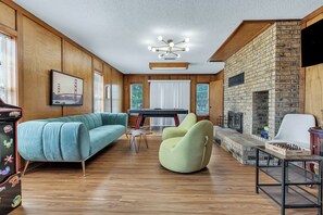 A spacious living room with a wooden floor, a blue sofa, a green armchair, a pool table, and a fireplace. - Southside: Swimming Pool/Air Hockey/PacMan/5BRs (College Station, TX)