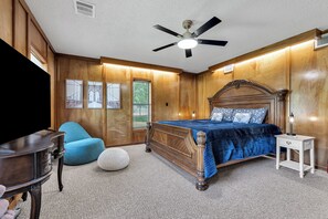 Une chambre à coucher avec un lit en bois, un ventilateur de plafond, une table de chevet, une lampe, une chaise bleue et un téléviseur fixé au mur.