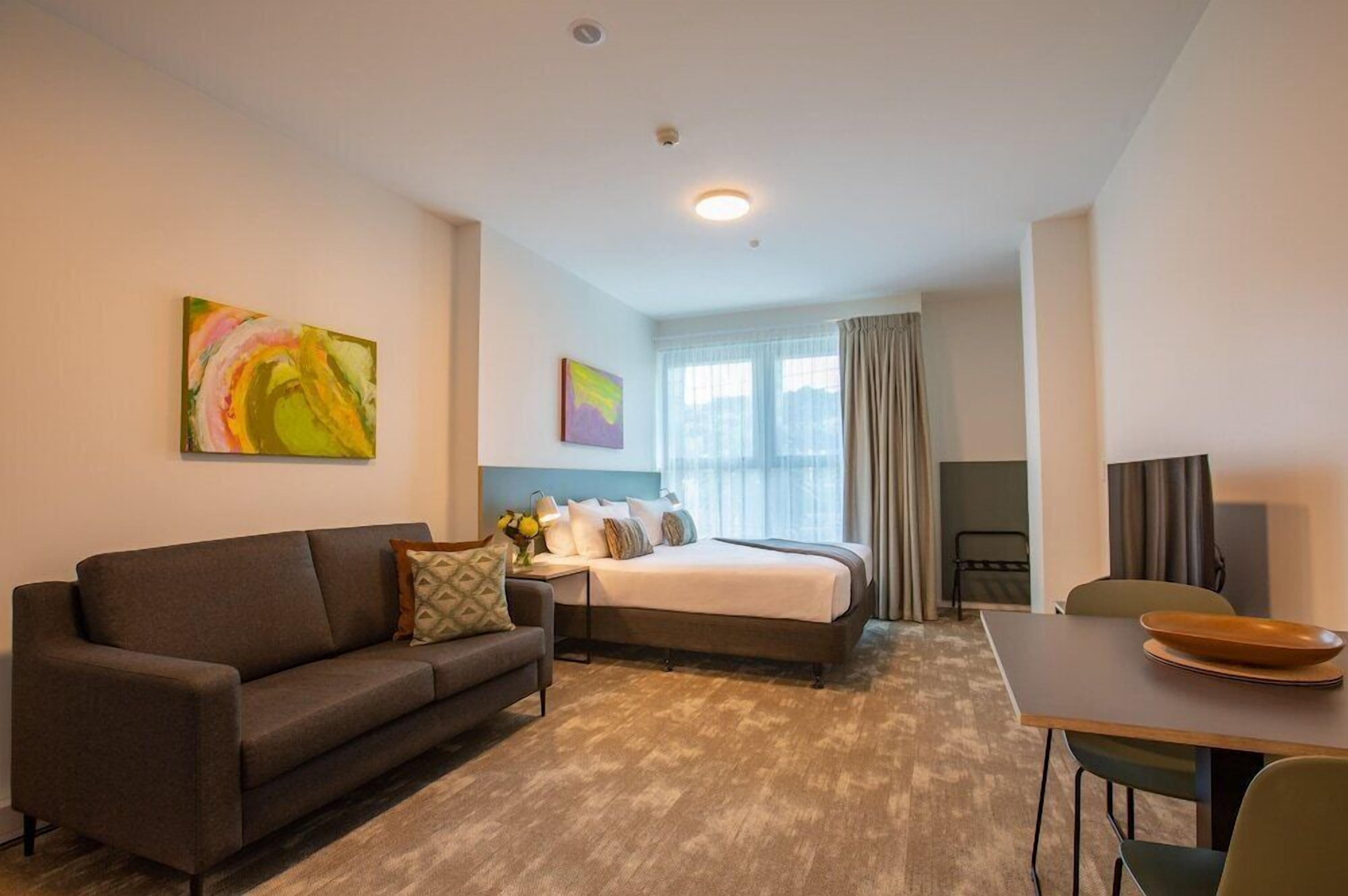 A hotel room with a bed, a sofa, a dining table, and artwork on the walls.