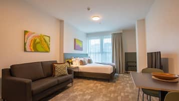 A hotel room with a bed, a sofa, a dining table, and artwork on the walls.