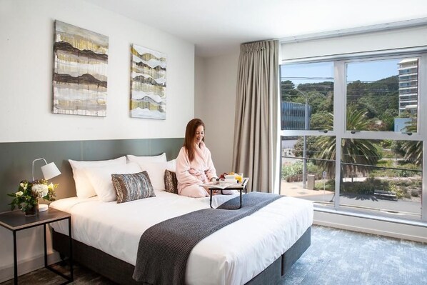 A hotel room with a large bed, a nightstand with a lamp, and a view of the outdoors through a large window. - Sojourn Apartment Hotel Riddiford Street Limited (Wellington)