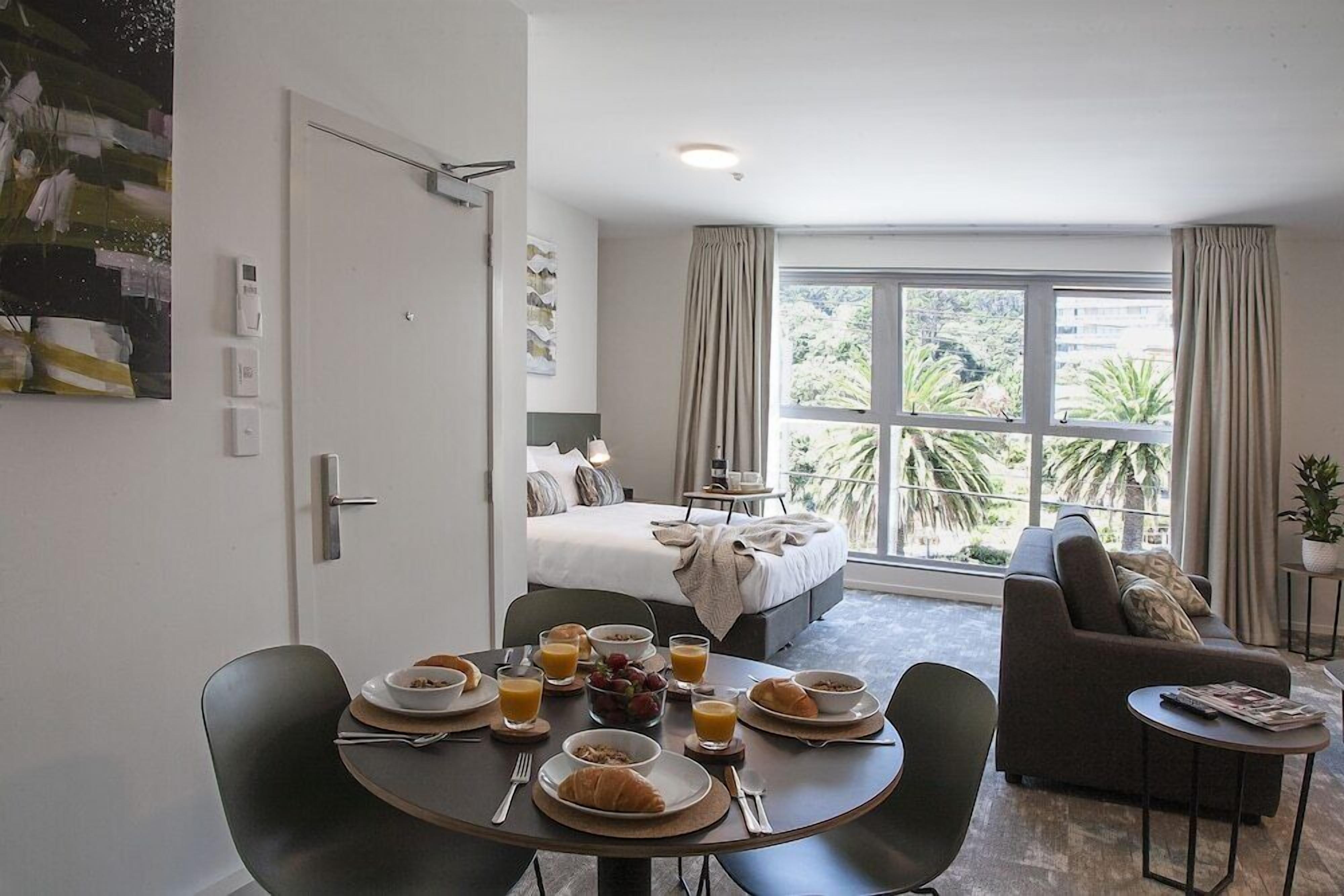 A hotel room with a bed, a dining table with breakfast, a sofa, and a large window with a view of greenery.