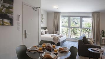 A hotel room with a bed, a dining table with breakfast, a sofa, and a large window with a view of greenery.