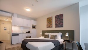 A compact hotel room with a bed, bedside tables, a desk, a chair, a kitchenette, and artwork on the walls.