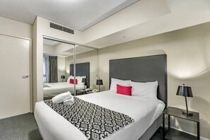 A hotel room with a large bed, a bedside table with a lamp, a mirror, and a door leading to another room. - Apartment 1112 ‘The middle of everywhere’ (Southbank)