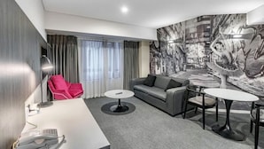 Una habitación de hotel moderna con un sofá gris, una silla roja, una mesita y un mural abstracto grande en la pared.