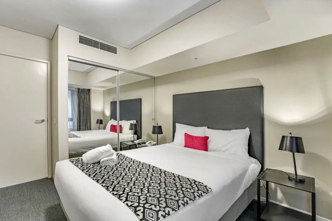 A hotel room with a large bed, a bedside table with a lamp, a mirror, and a door leading to another room.
