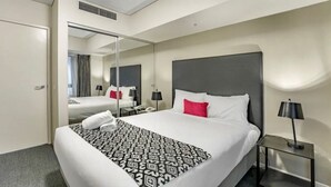 Habitación de hotel con una cama grande, una mesita de noche con lámpara, un espejo y una puerta que da a otra habitación.