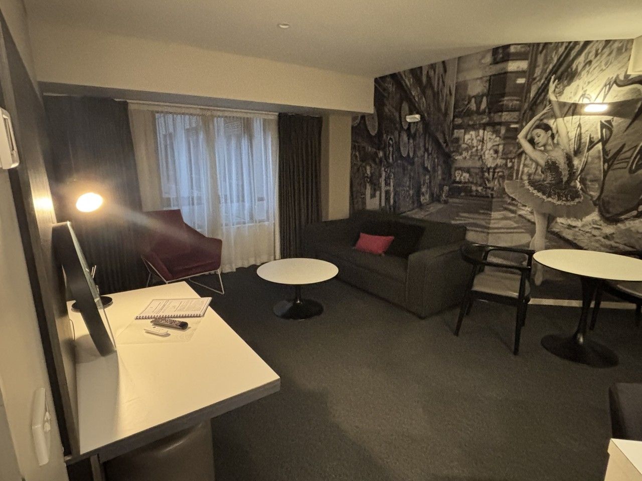 A modern hotel room with a flat-screen TV, a desk with a notebook and pen, a round dining table, a sofa with a red pillow, and a large abstract mural on the wall.