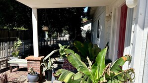 A covered porch with potted plants, a red chair, and a white building in the background.