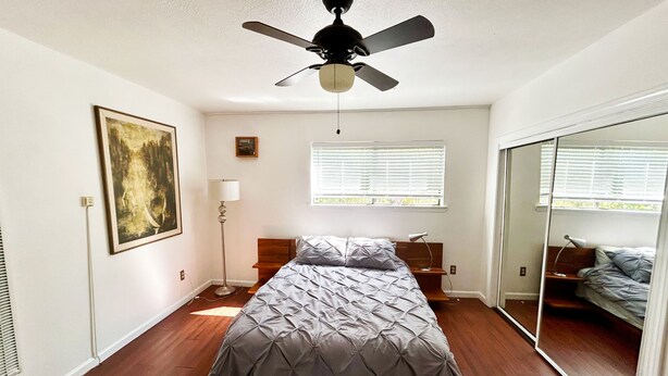 A bedroom with a bed, a ceiling fan, a window with blinds, a floor lamp, and a painting on the wall.