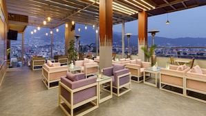 A rooftop lounge area with seating and tables, large windows offering a city view, and a wooden ceiling with lighting fixtures. - Regenta Place Sabrina Kathmandu (Burhanilkantha)