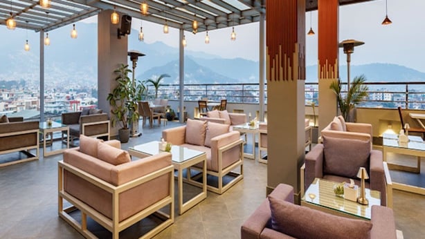 A rooftop lounge with sofas and tables, large windows offering a city view, and hanging pendant lights. - Regenta Place Sabrina Kathmandu (Burhanilkantha)
