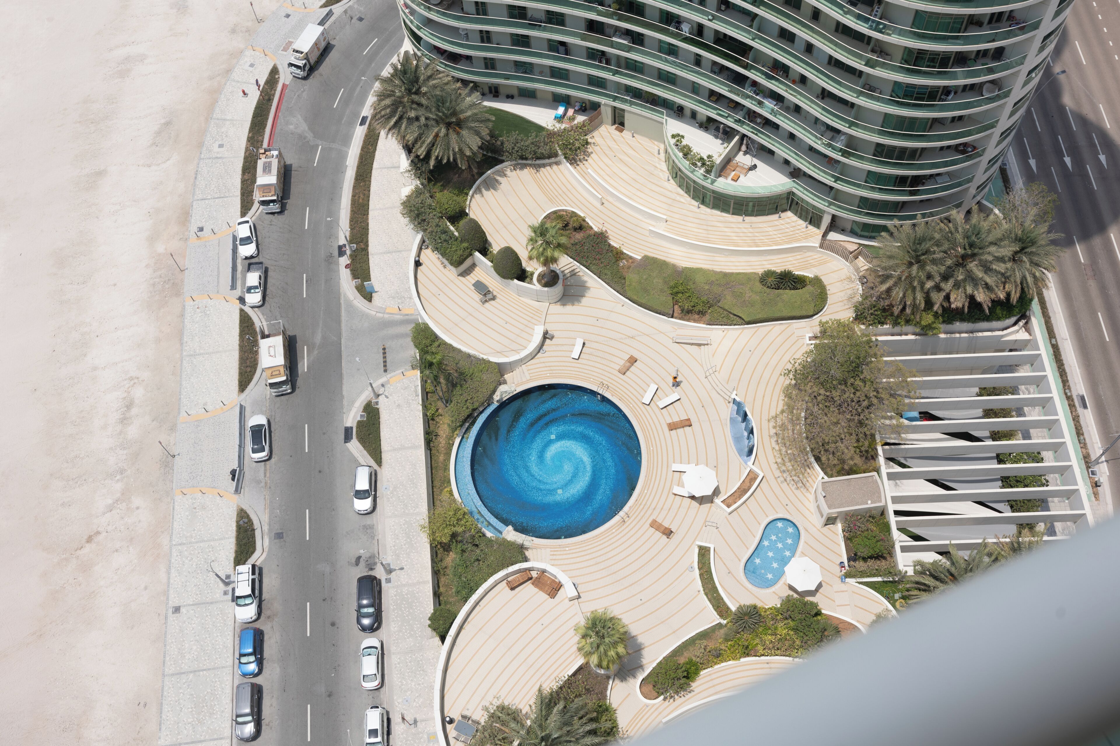 An aerial view of a modern hotel with a central swimming pool, landscaped areas, and a curved road with parked cars.