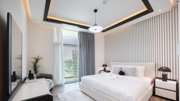 A modern hotel room with a large bed, bedside tables, a desk, and a view of the city through the window. - Bloomfields 3BR plus Maid in Beach Tower (Abu Dhabi)