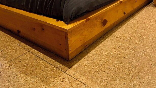 Una cama individual con estructura de madera y ropa de cama oscura en una habitación con suelo de baldosas y una cortina azul.