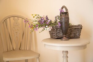 Una silla con un respaldo decorativo, una cesta de mimbre y un florero metálico con flores sobre una mesita redonda.