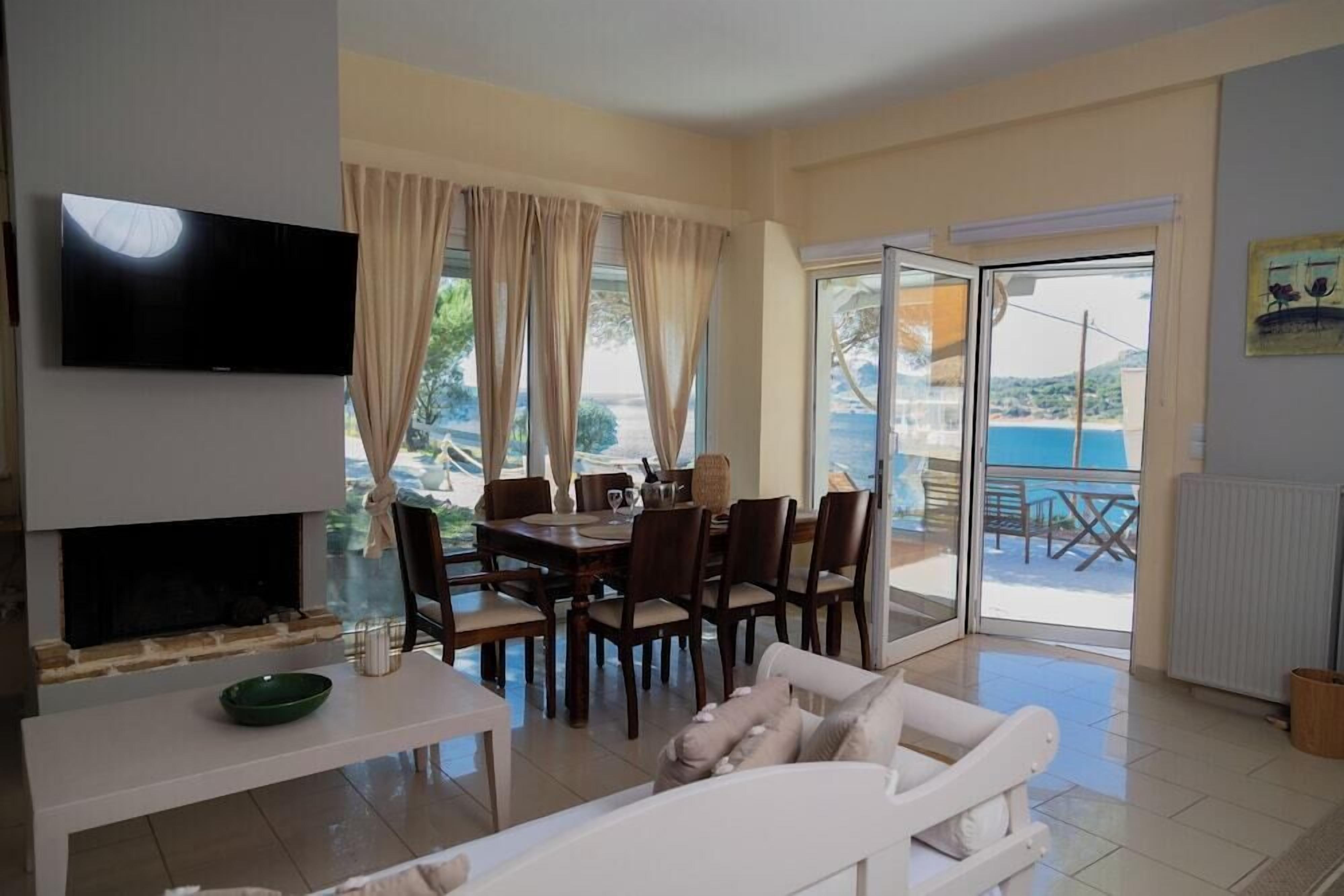 A living room with a TV, dining table, chairs, and a sofa. There is a sliding glass door leading to an outdoor area with a view of the sea.