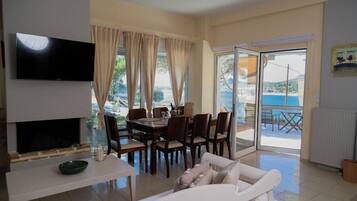 A living room with a TV, dining table, chairs, and a sofa. There is a sliding glass door leading to an outdoor area with a view of the sea.