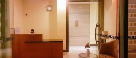 A reception area with a reception desk, a sofa, a small table with a vase, and a door leading to another room.