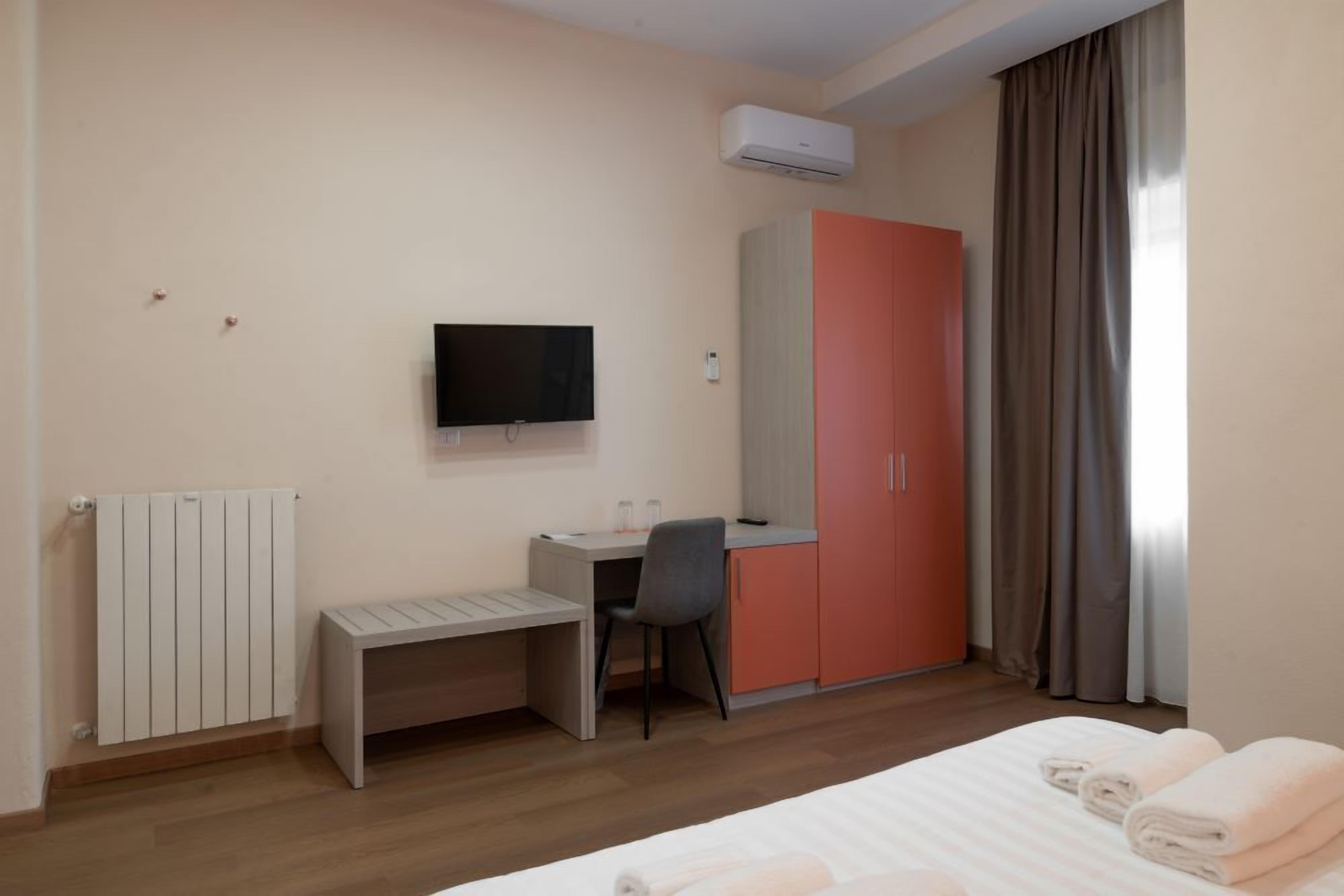 Habitación de hotel con cama, escritorio, silla, televisión, armario y radiador.