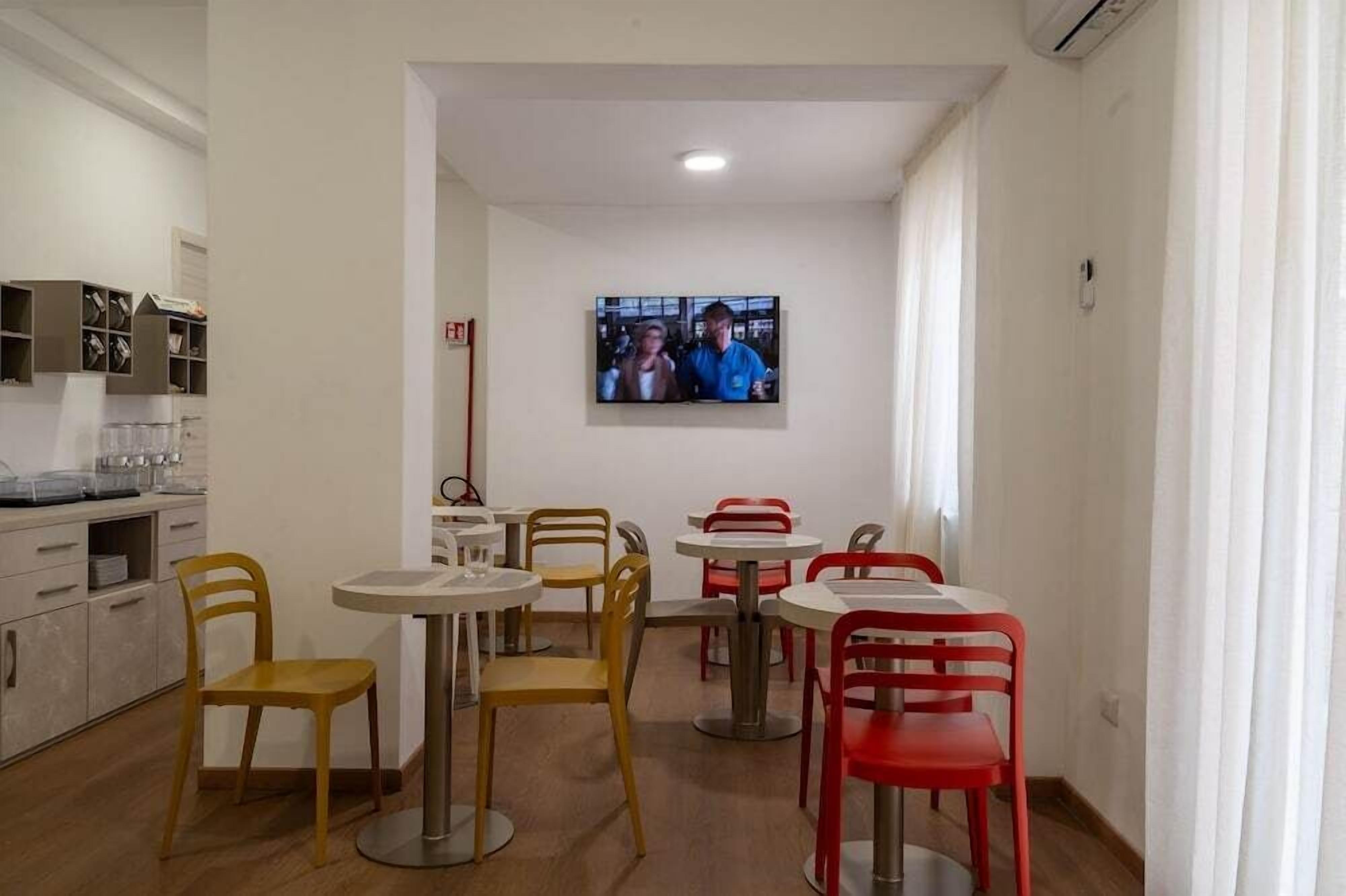 A dining area with multiple tables and chairs, a television mounted on the wall, and a kitchen area with cabinets and a countertop.