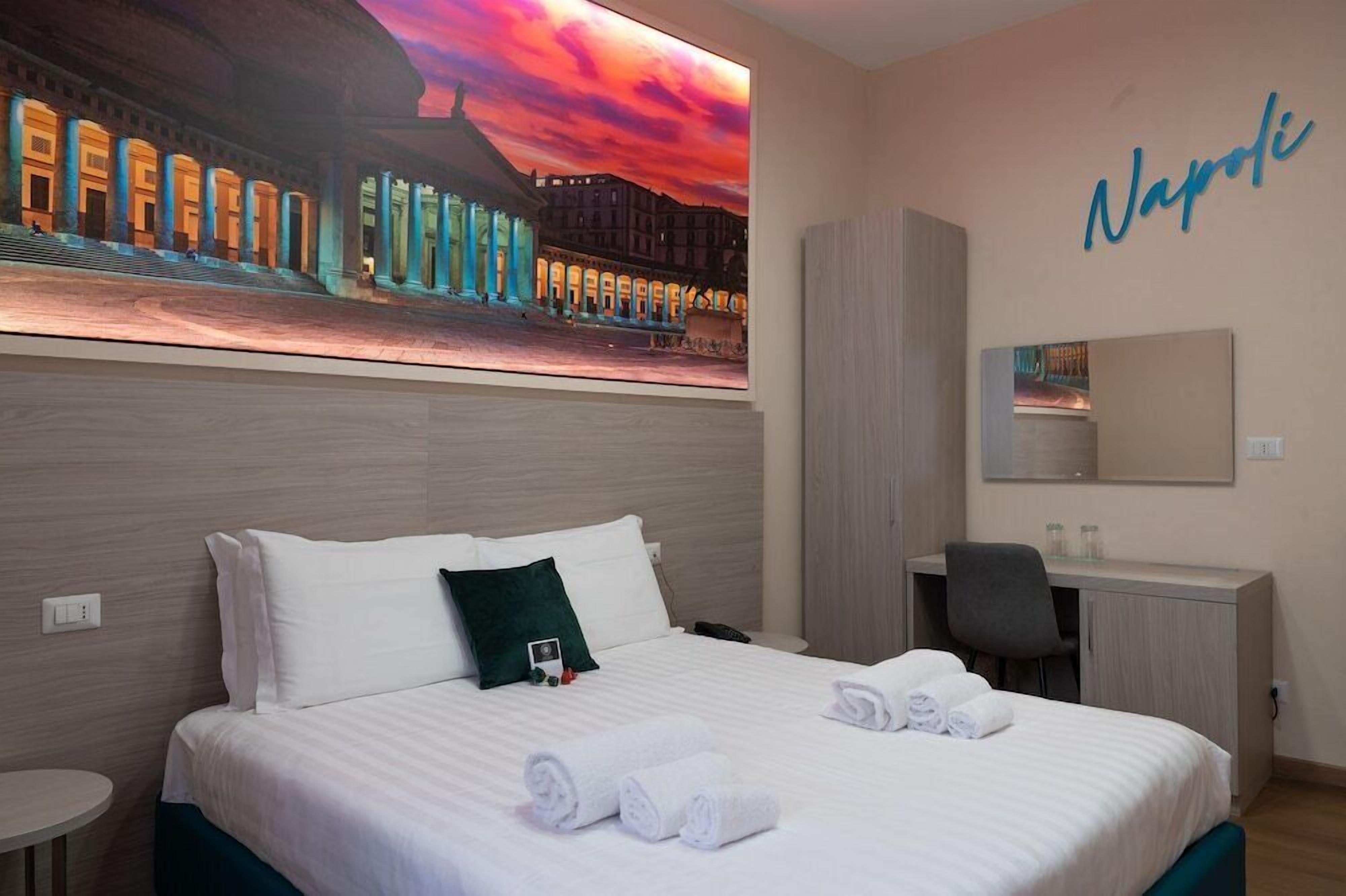 A hotel room with a bed, a desk, a chair, a small table, and a large wall-mounted artwork.