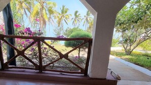 Un espacio al aire libre techado con piso de baldosas, barandilla de madera y vistas a palmeras y flores.