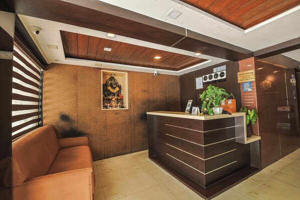 A reception area with a wooden reception desk, a seating area with a sofa, and a wall-mounted decorative item. - Hotel Maruya Palace (Mysore)