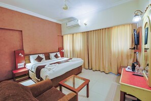 A hotel room with a bed, a desk, a chair, a mirror, and a television. - Hotel Maruya Palace (Mysore)