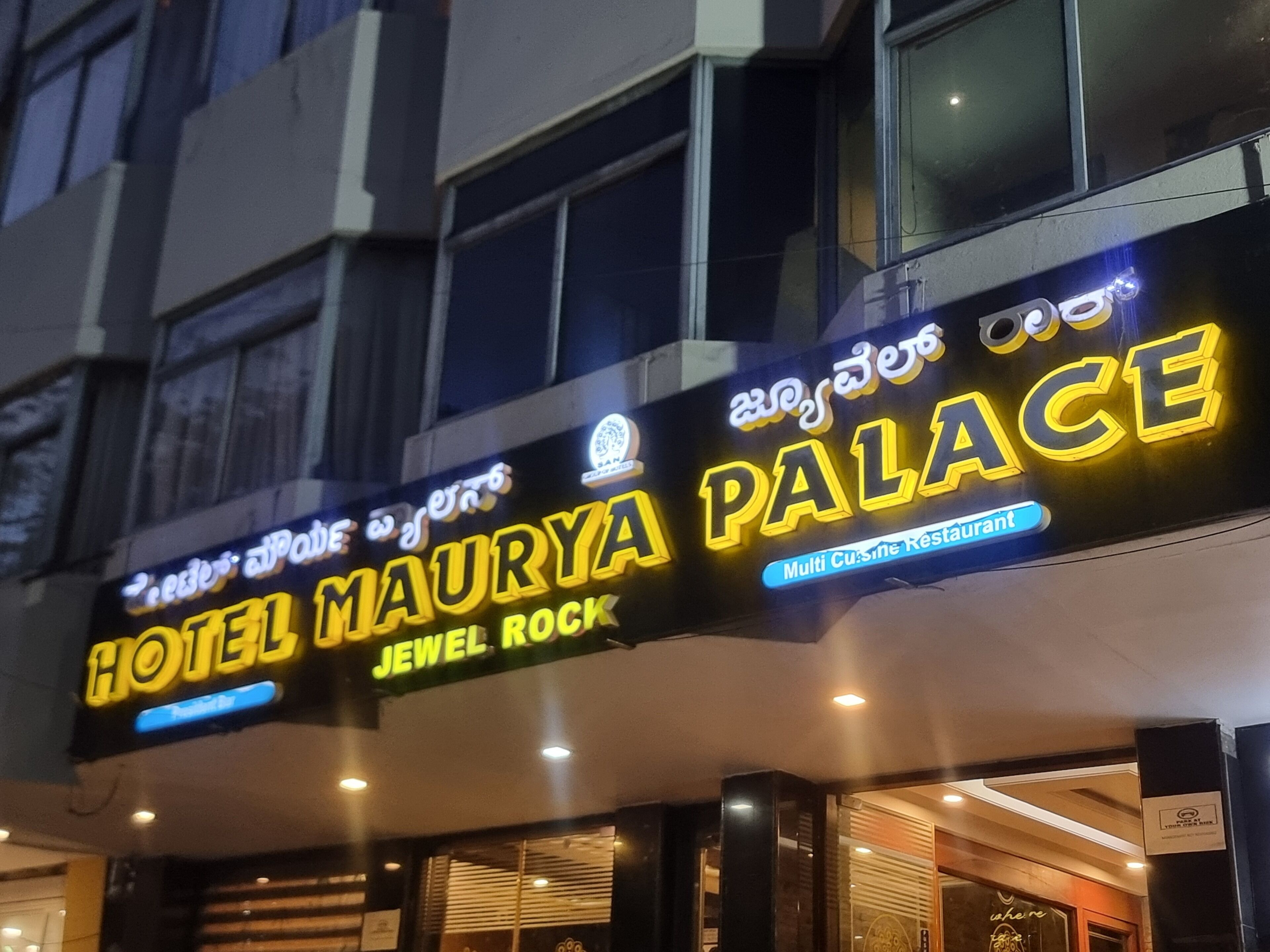 Placa de hotel com 'HOTEL MAURY PALACE JEWEL ROCK' e 'Restaurante com Vários Tipos de Cozinha' escrita em letras brilhantes e iluminadas.