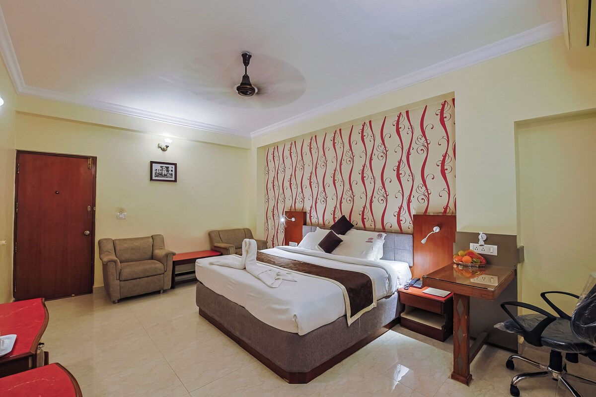 A hotel room with a bed, two armchairs, a desk with a chair, a ceiling fan, and a wall with a decorative pattern.