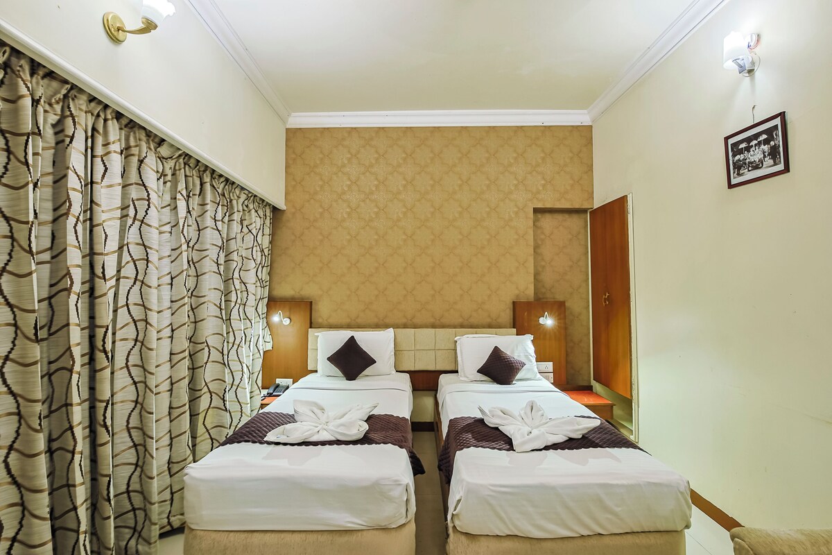 A hotel room with two single beds, a patterned curtain, a wall-mounted light fixture, and a framed photo on the wall.