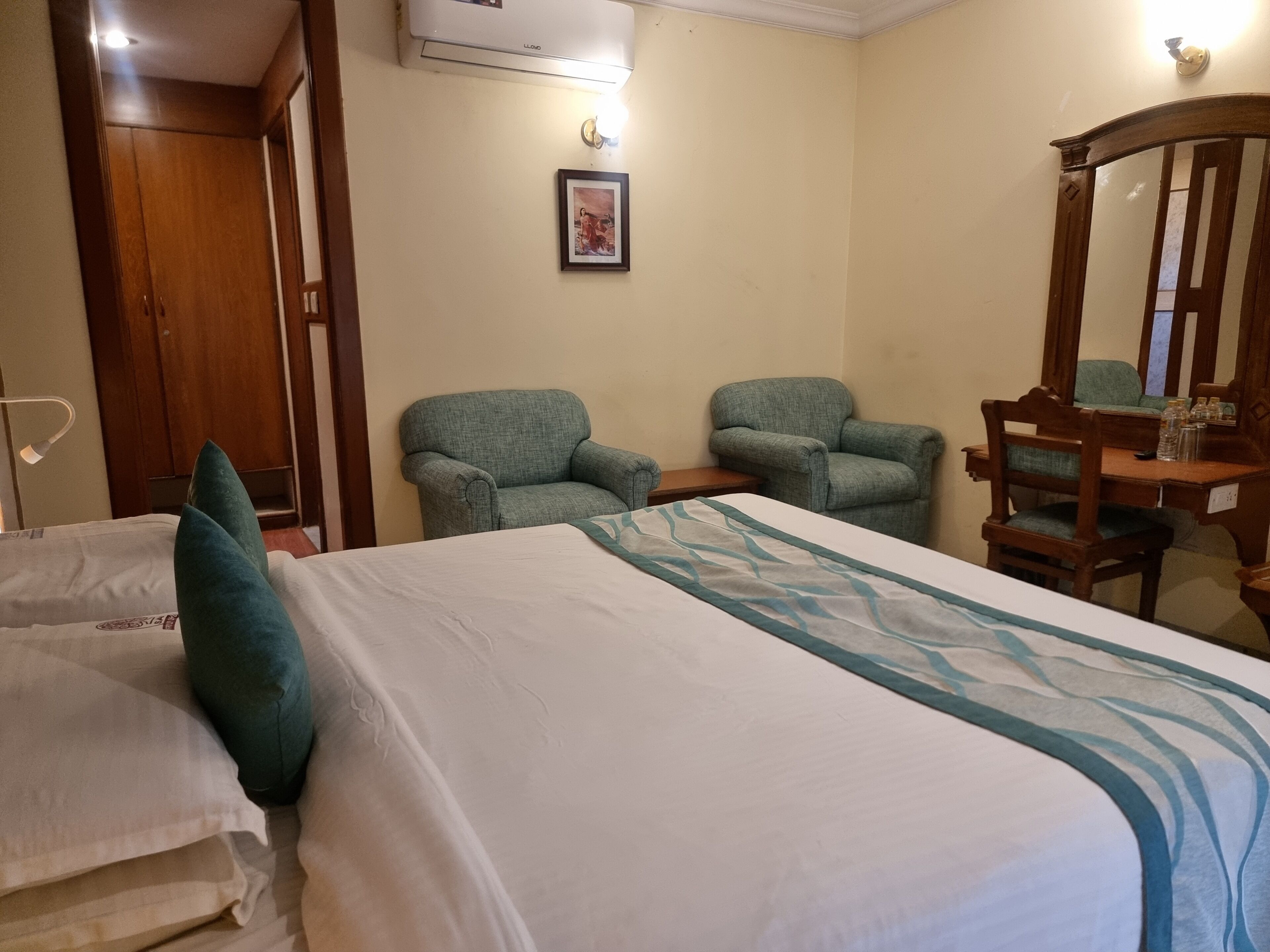 Habitación de hotel con cama, dos sillones, una silla de madera, un espejo y un equipo de aire acondicionado.