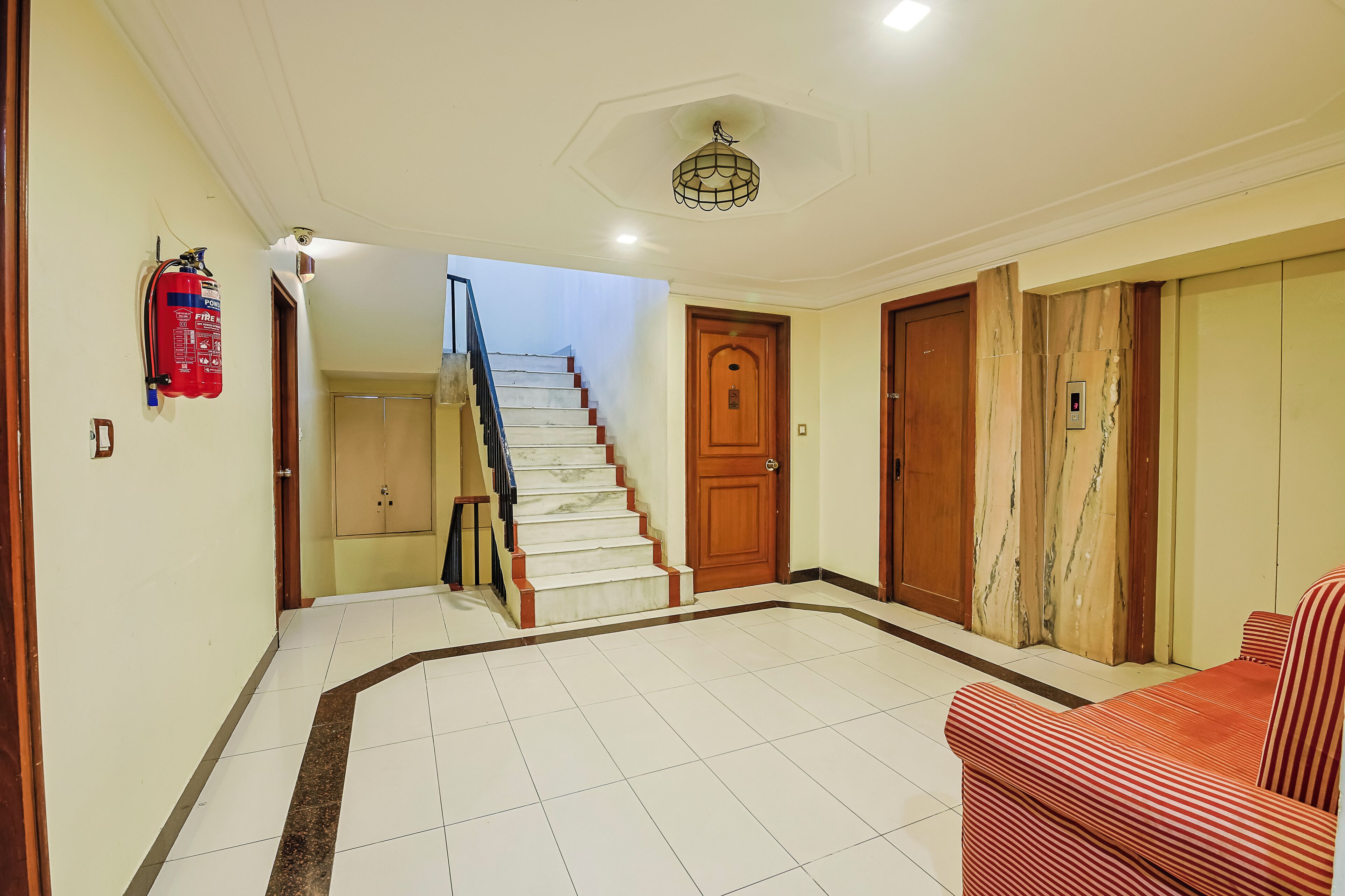 Un vestíbulo de hotel con una escalera, un extintor, una puerta con pantalla digital y un sofá estampado.