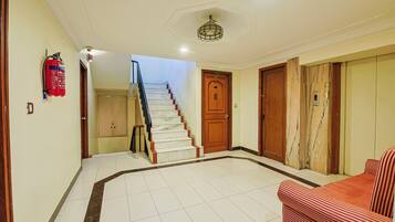 A hotel lobby with a staircase, a fire extinguisher, a door with a digital display, and a patterned sofa.