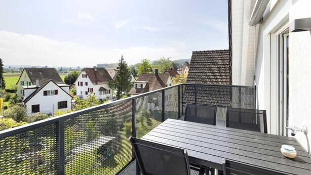 A balcony with a table and chairs, overlooking a residential area with houses and greenery.