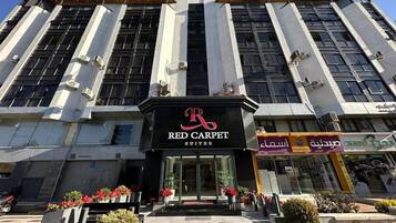 Un edificio con un llamativo letrero que dice 'Red Carpet Suites', una farmacia y otra farmacia con rótulos en árabe.