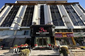 En bygning med et tydeligt skilt, der viser 'Red Carpet Suites', samt en apotek og et apotek med arabisk skiltemateriale.