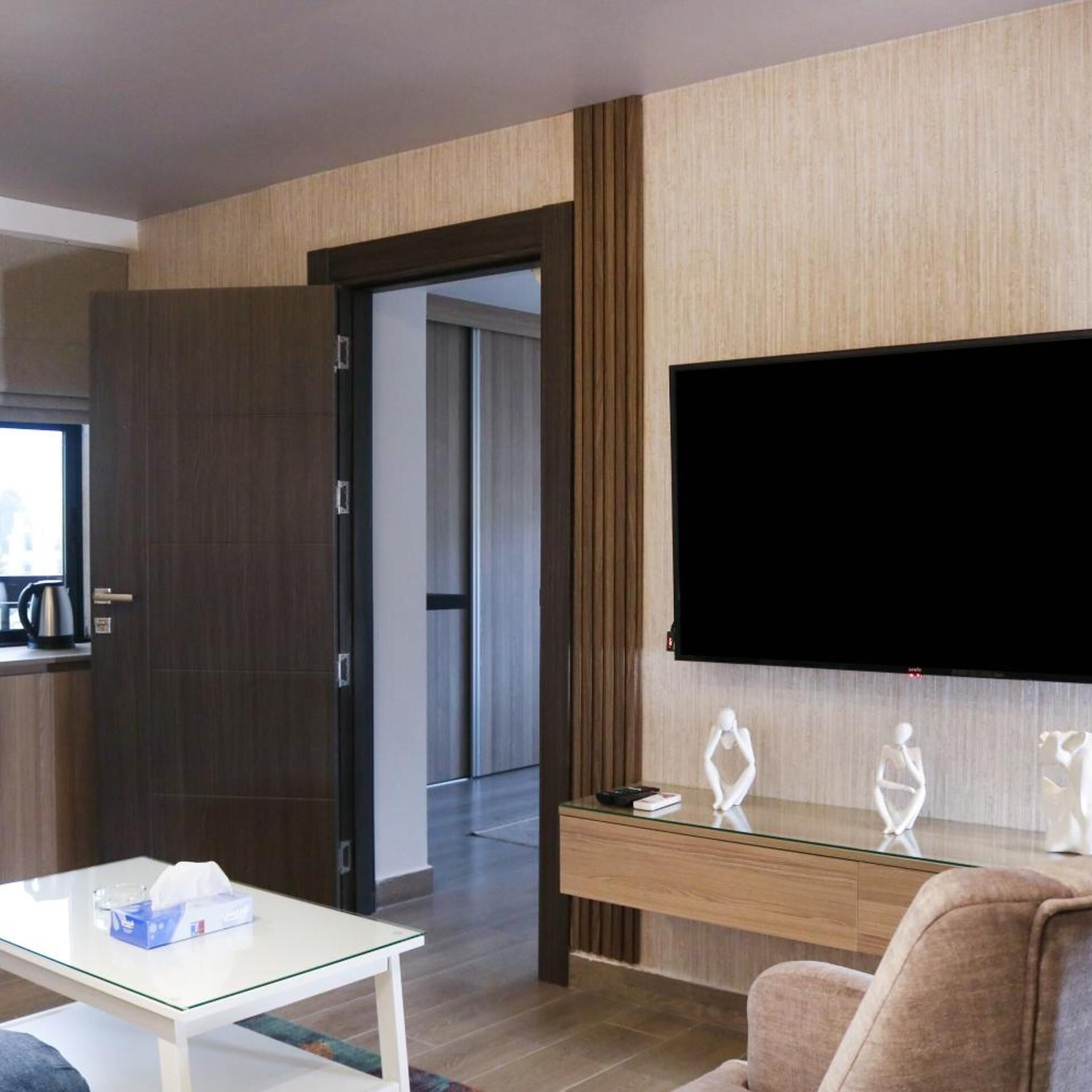 A modern hotel room with a flat-screen TV mounted on the wall, a glass-top coffee table, a plush armchair, and a wooden console table with decorative items.