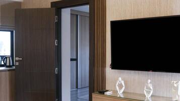 A modern hotel room with a flat-screen TV mounted on the wall, a glass-top coffee table, a plush armchair, and a wooden console table with decorative items.