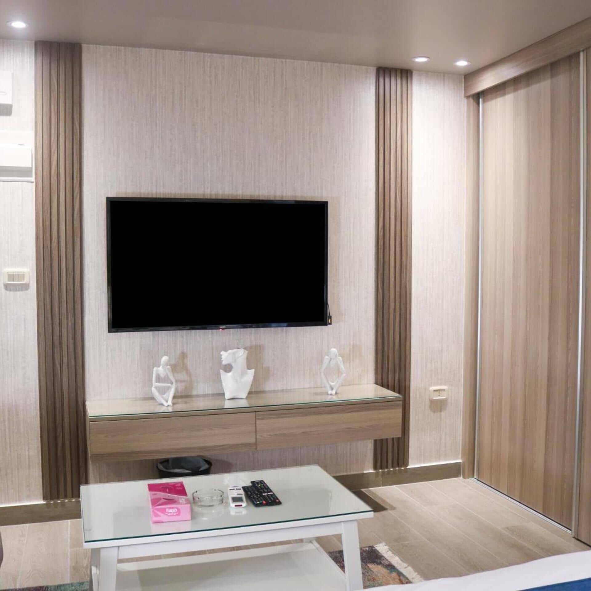 Una habitación de hotel moderna con un televisor de pantalla plana montado en la pared, una mesa de centro de vidrio con un control remoto, un teléfono inteligente y un pequeño objeto decorativo.