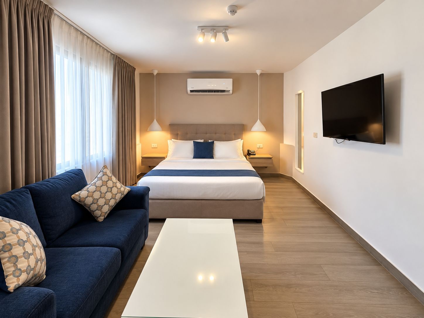 A hotel room with a bed, a sofa, a TV, and a ceiling-mounted air conditioning unit.