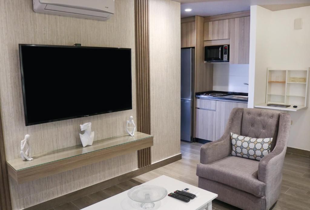 A modern living room with a flat-screen TV, a glass coffee table, a comfortable armchair, and a kitchen area in the background.