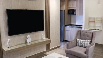 A modern living room with a flat-screen TV, a glass coffee table, a comfortable armchair, and a kitchen area in the background.