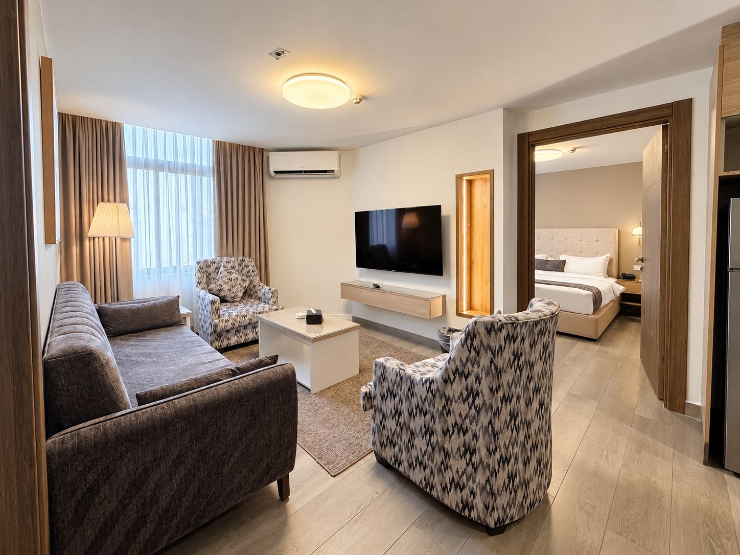 Une chambre d’hôtel moderne équipée d’un canapé, d’un fauteuil, d’une télévision, d’une table de chevet et d’un lit avec une tête de lit.
