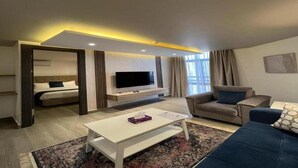 A hotel room with a bed, a TV mounted on the wall, a sofa, a coffee table, and a patterned rug. - Red Carpet Hotel & Suite (Amman)