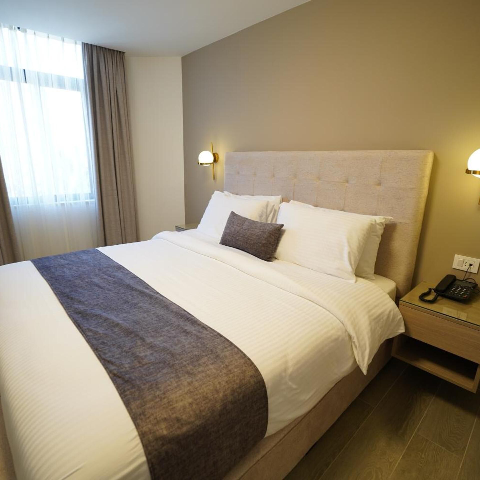 Ein Hotelzimmer mit einem großen Bett, einem Nachttischtelefon und einem fensterbelaubten Fenster.