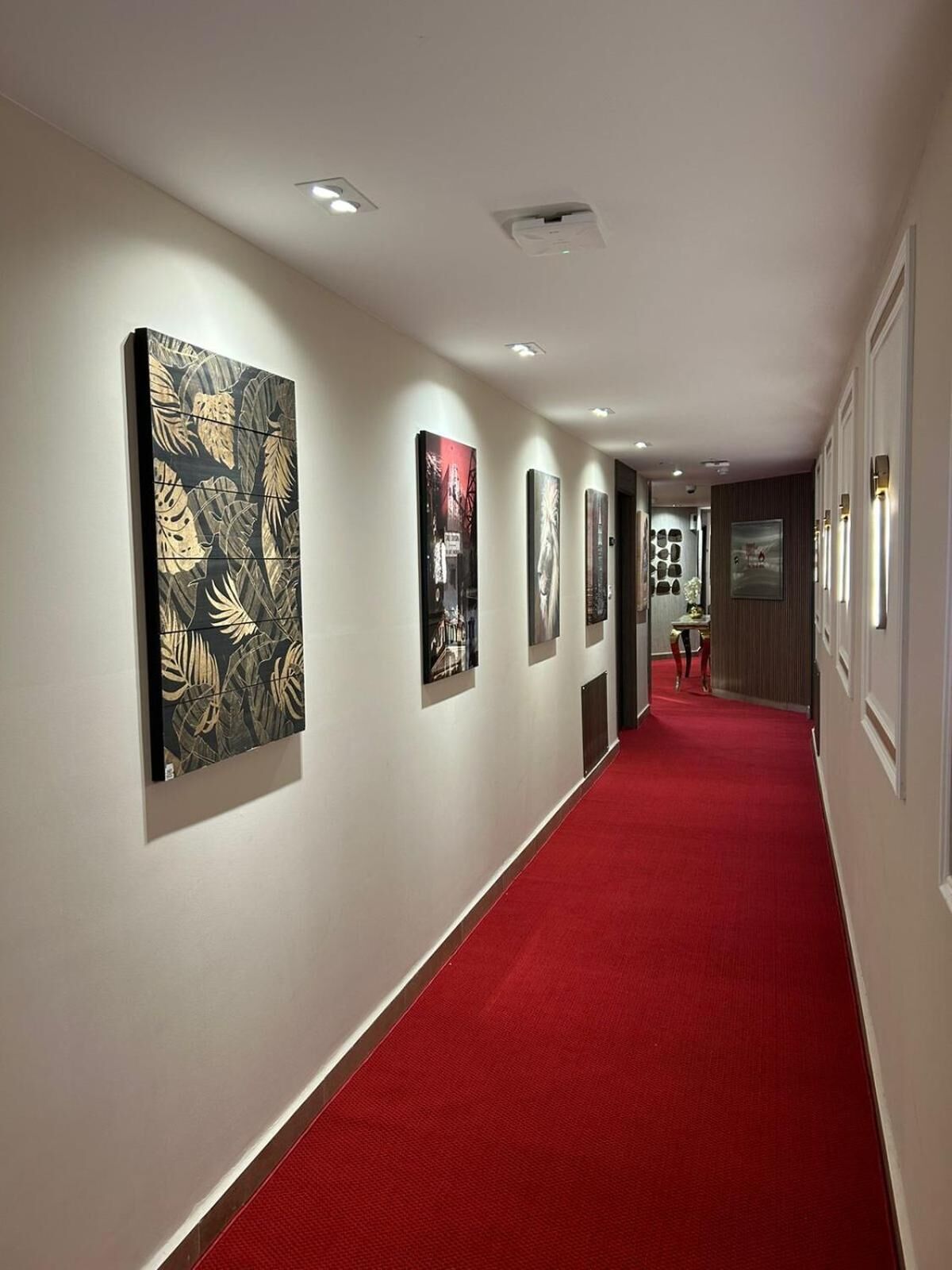 Un pasillo con alfombra roja, paredes blancas y obras de arte a la izquierda.
